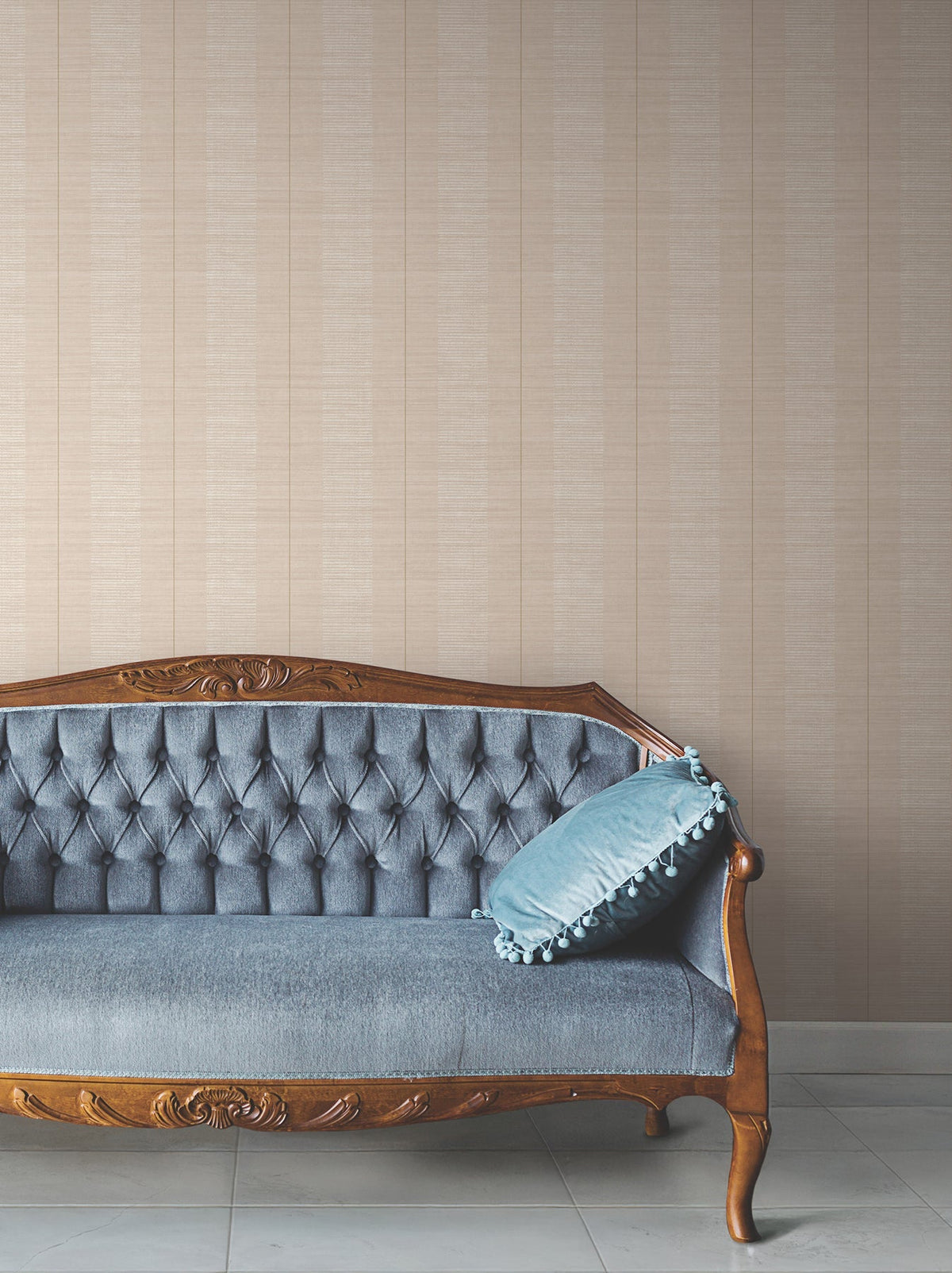 A vintage wooden sofa with blue tufted upholstery and a blue pillow sits against a wall adorned with Colbert Stripe Textured Wallpaper on a tiled floor.