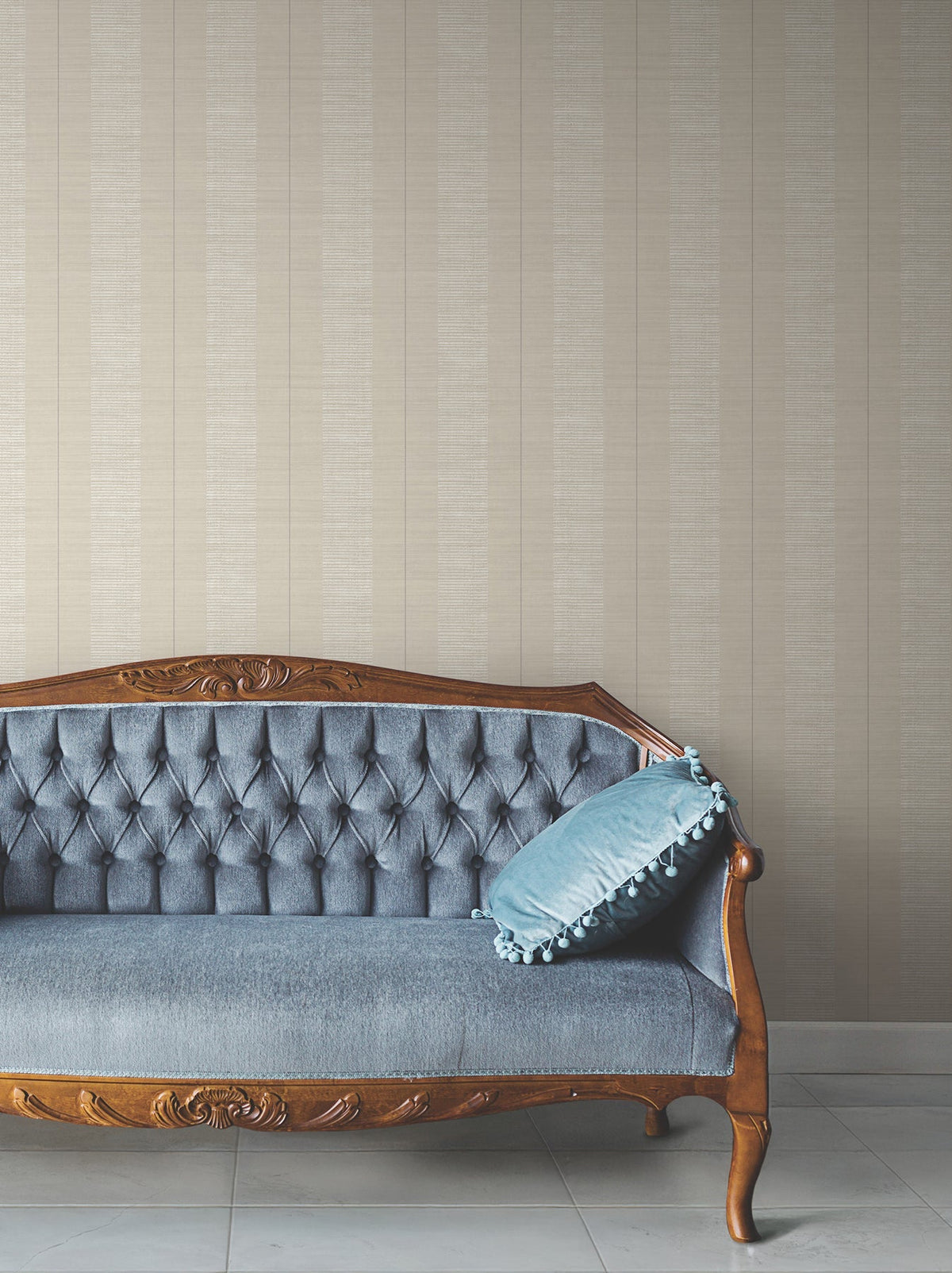 A vintage blue tufted sofa with a carved wooden frame sits against a wall adorned with Colbert Stripe Textured Wallpaper. A blue cushion with pompom trim is placed on the right side of the sofa.