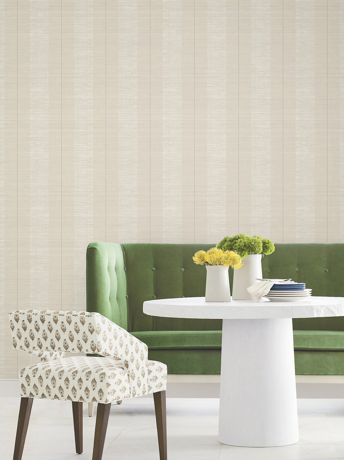 Modern dining area featuring a round white table, green tufted bench, patterned white chair, and vases of yellow and green flowers, set against a wall adorned with Colbert Stripe Textured Wallpaper.