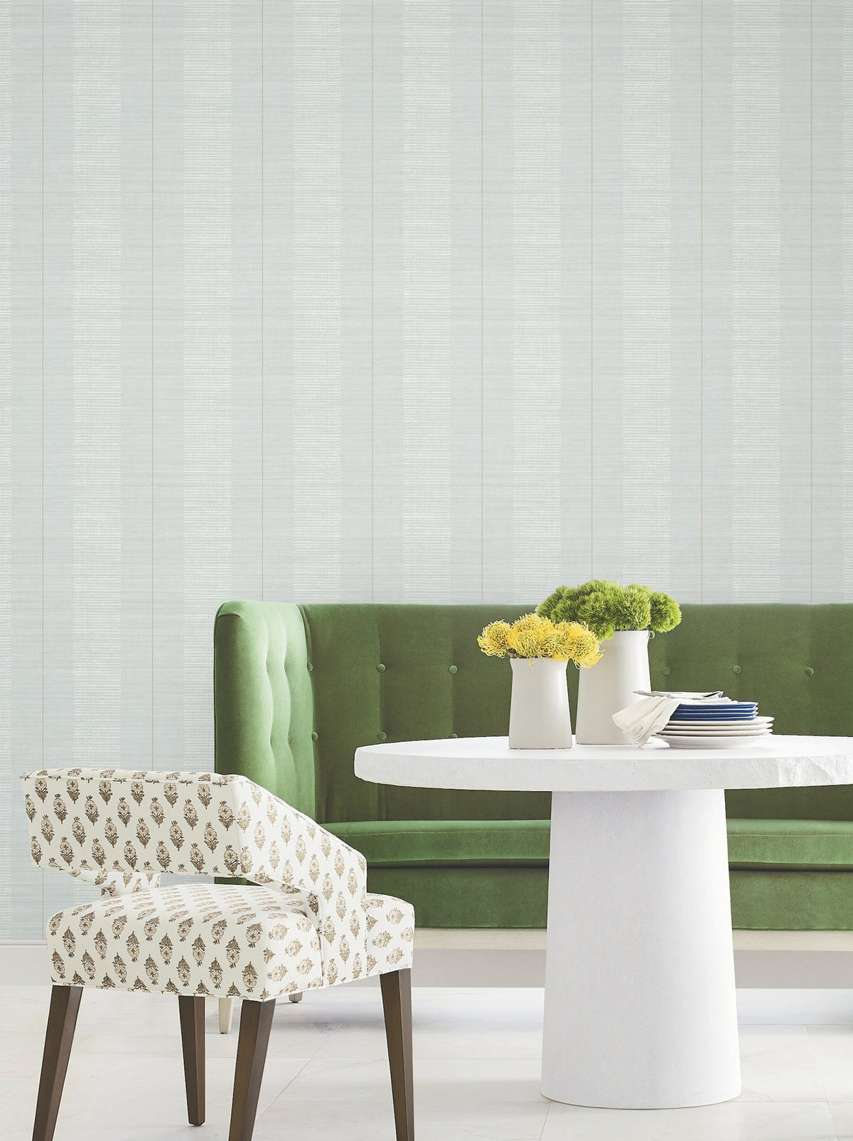 Modern dining area featuring a green tufted sofa, a white round table adorned with yellow and green flowers, and a patterned white chair on a light floor, set against Colbert Stripe Textured Wallpaper.