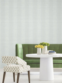 Modern dining area featuring a green tufted sofa, a white round table adorned with yellow and green flowers, and a patterned white chair on a light floor, set against Colbert Stripe Textured Wallpaper.