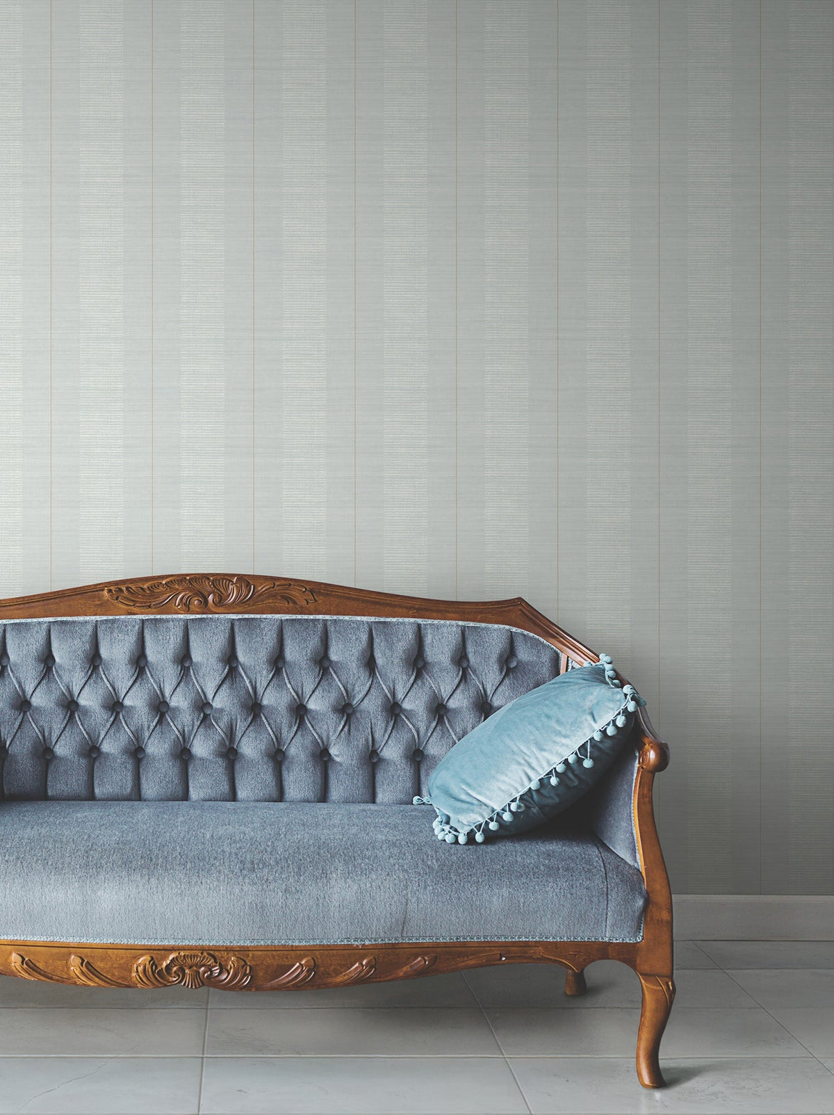 A vintage wooden sofa with blue tufted upholstery and a blue cushion sits against a tiled floor, backed by a light gray wall adorned with Colbert Stripe Textured Wallpaper.