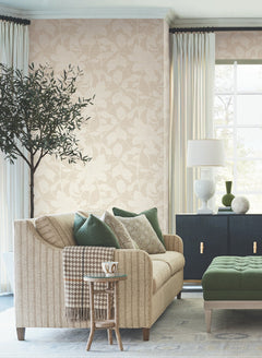 A cozy, sunlit living room features Solange Botanical Wallpaper by York Wallcoverings as a backdrop for a beige sofa adorned with green and patterned pillows next to a side table holding a mug. An olive tree, green ottoman, black cabinet with a white lamp, and large window with light curtains complete the inviting space.