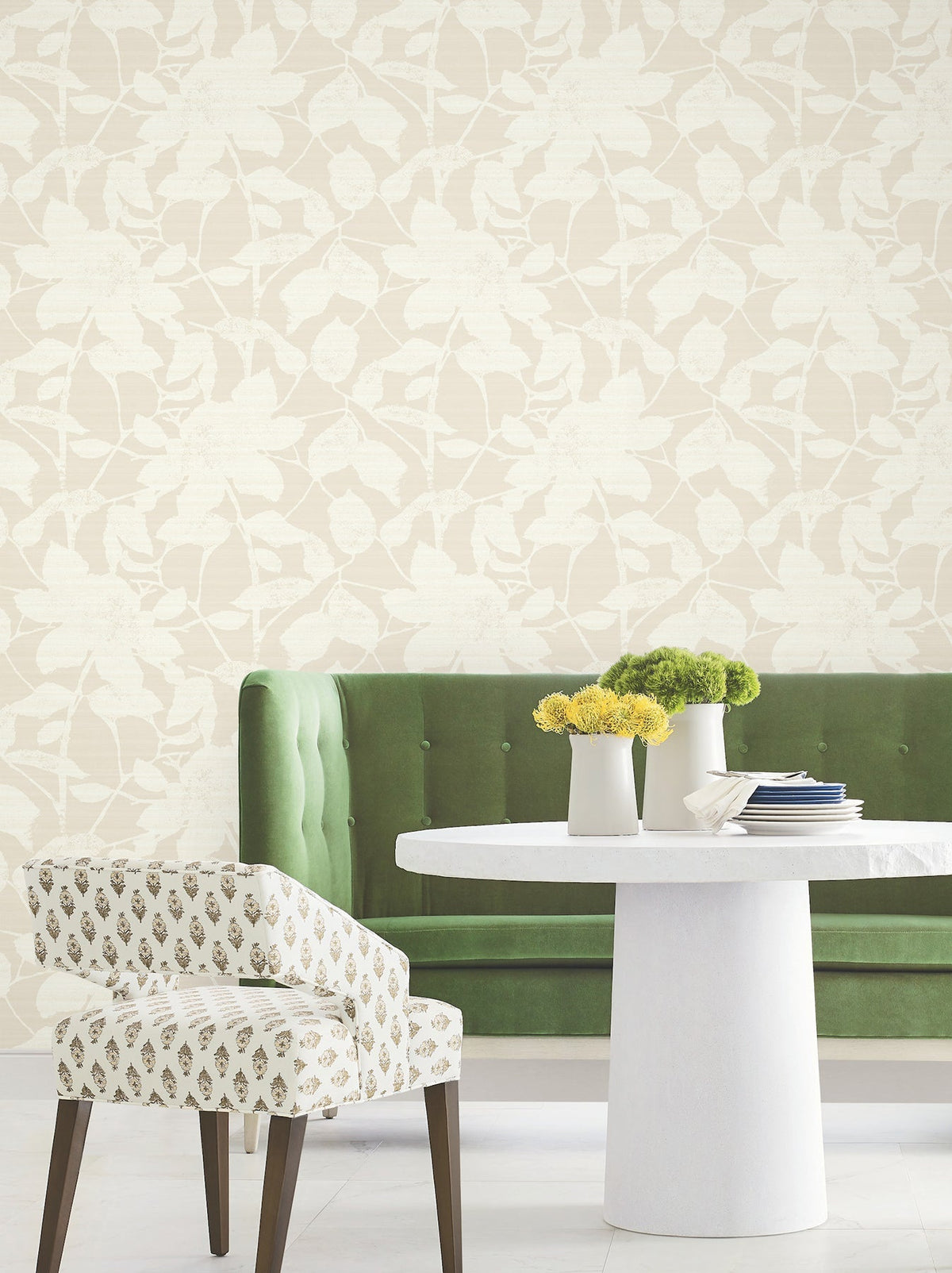 A green tufted sofa, a round white table with small vases of yellow flowers, and a white patterned chair sit in front of the Solange Botanical Wallpaper by York Wallcoverings.