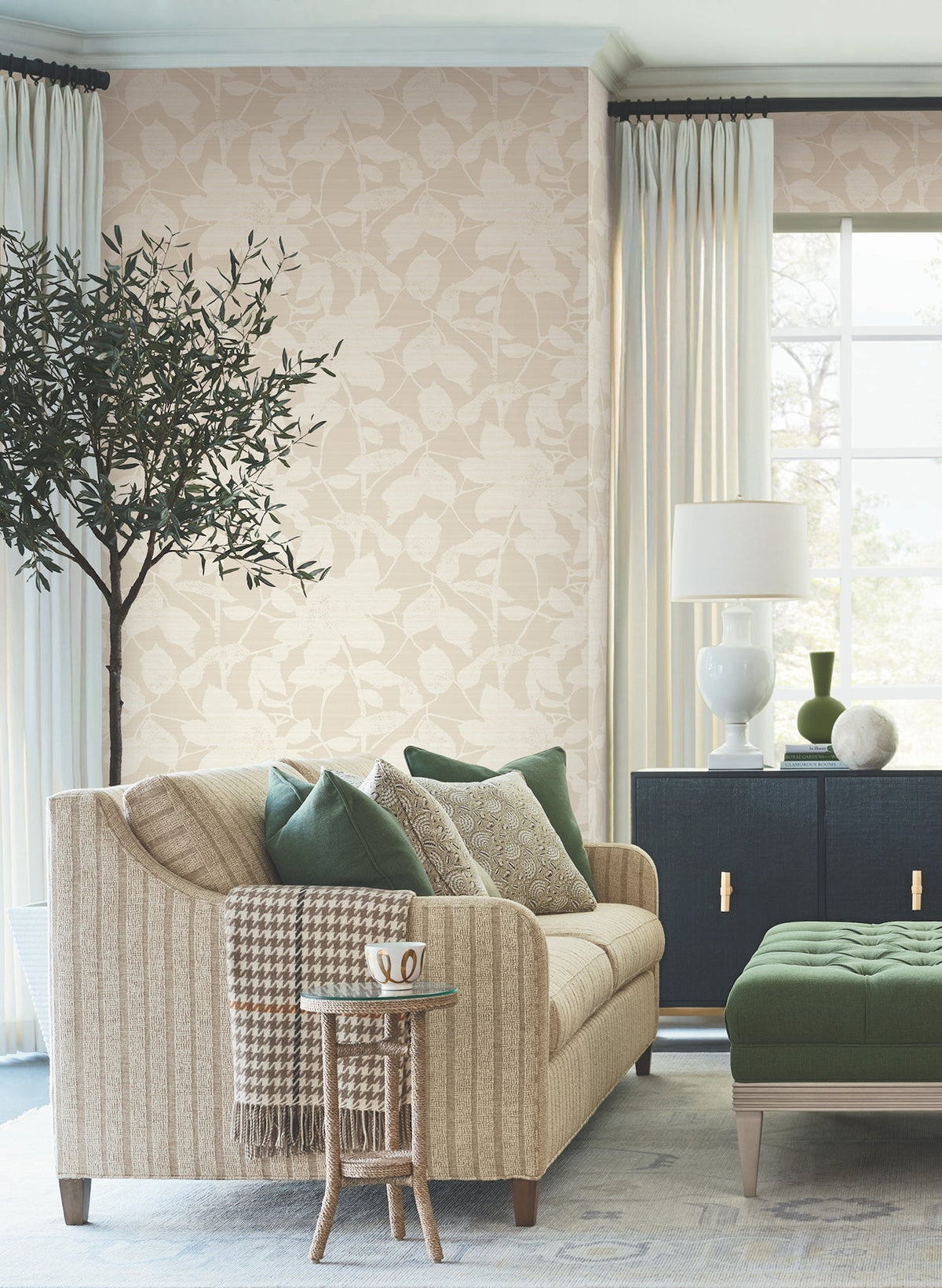 A cozy living room featuring York Wallcoverings’ Solange Botanical Wallpaper, a beige sofa with patterned pillows, green accents, a potted tree, a side table with a mug, a black cabinet, a lamp, and sheer curtains on the window letting in natural light.