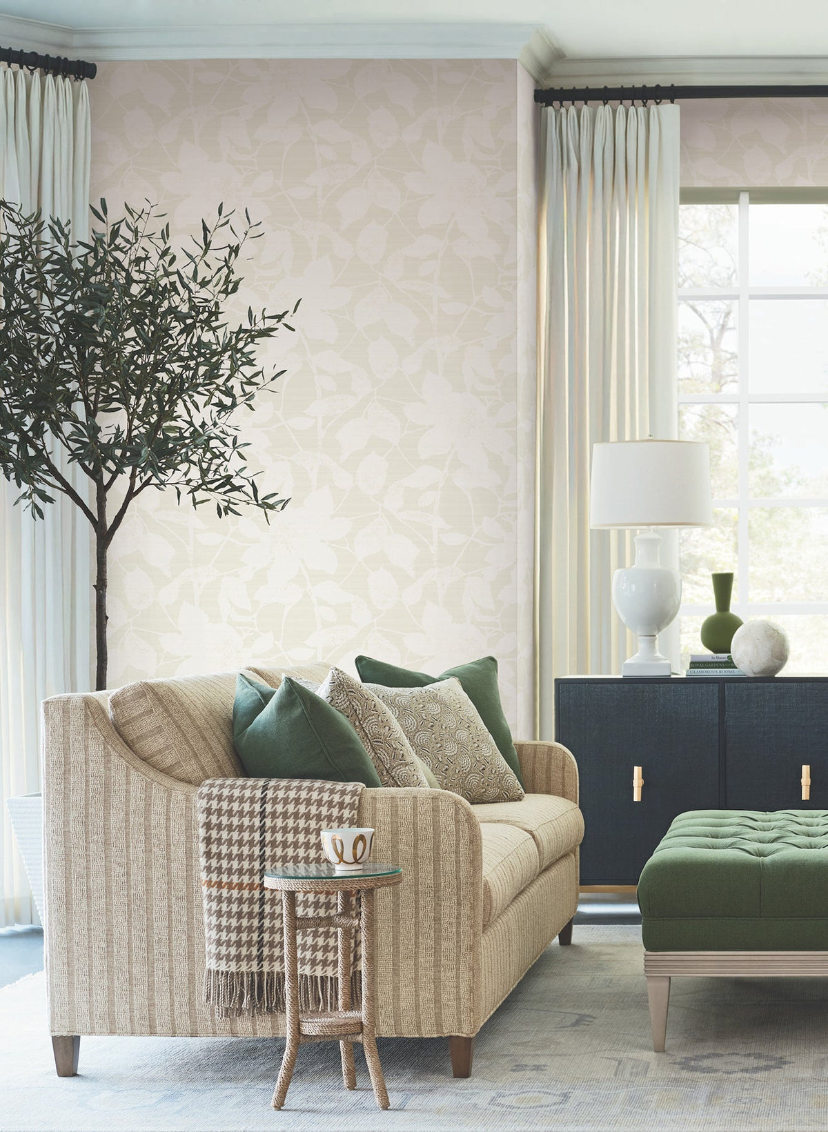 A cozy living room featuring Solange Botanical Wallpaper by York Wallcoverings, with a beige sofa, green and neutral pillows, a small round side table with a mug, an ottoman, a potted tree, a black cabinet, white lamp, and sheer curtains framing a bright window.