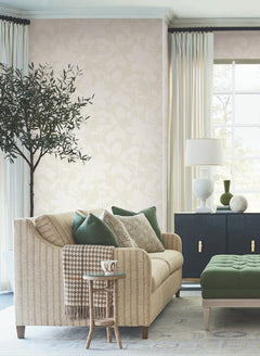 A cozy living room featuring Solange Botanical Wallpaper by York Wallcoverings, with a beige sofa, green and neutral pillows, a small round side table with a mug, an ottoman, a potted tree, a black cabinet, white lamp, and sheer curtains framing a bright window.