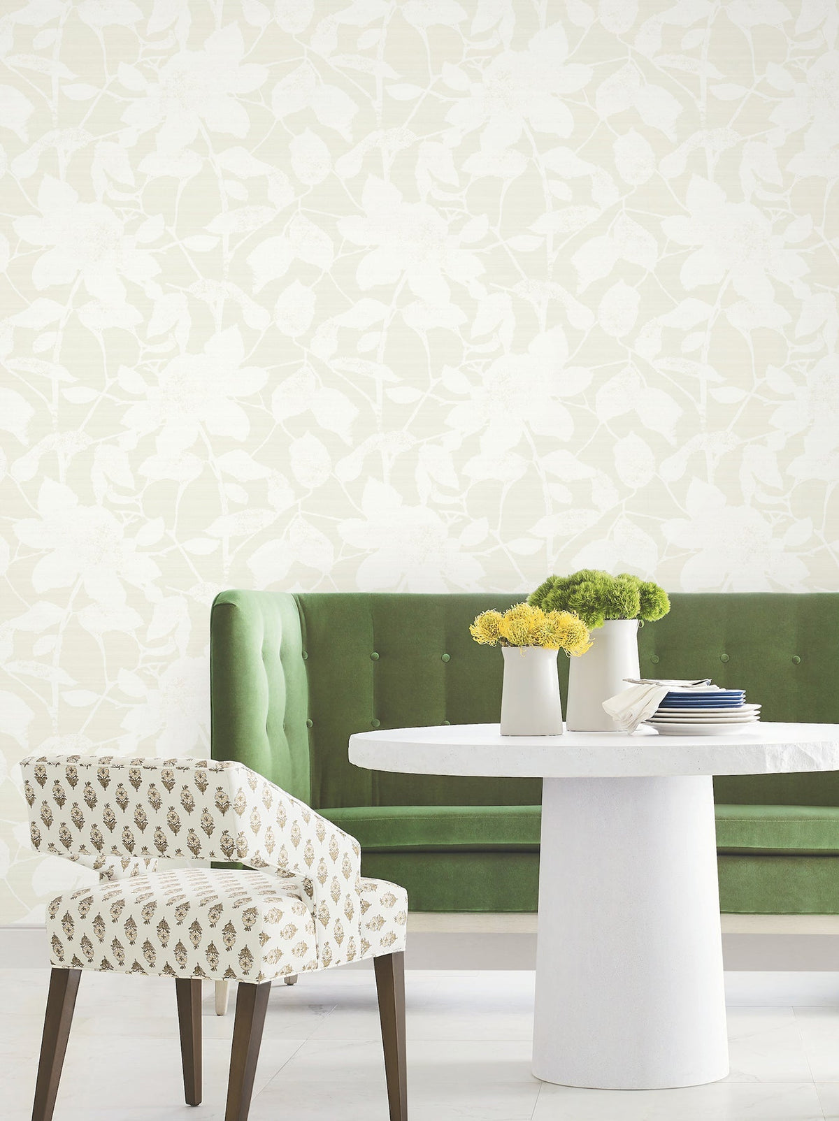 Green velvet tufted bench, white round table with white and yellow flowers in vases, patterned chair, and a Solange Botanical Wallpaper by York Wallcoverings as the background.