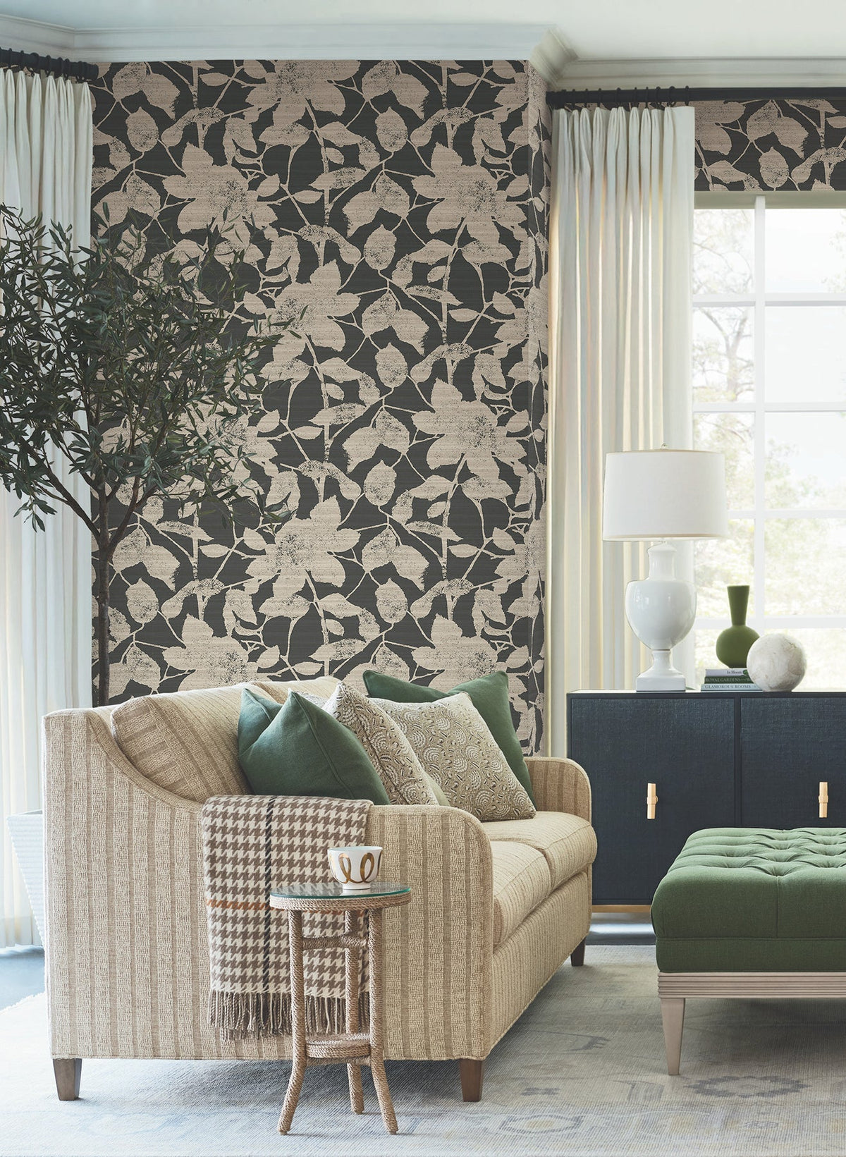 Living room featuring a striped tan sofa with green and neutral pillows, a side table holding a mug, green ottoman, white curtains, a potted plant, dark blue cabinet, and a white lamp near a sunny window—all set against Solange Botanical Wallpaper by York Wallcoverings.