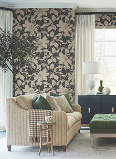 A cozy living room featuring a beige sofa with green and patterned pillows, a houndstooth throw, a small side table with a mug, and a green ottoman, set against an accent wall adorned with Solange Botanical Wallpaper by York Wallcoverings. Light curtains frame the large window.