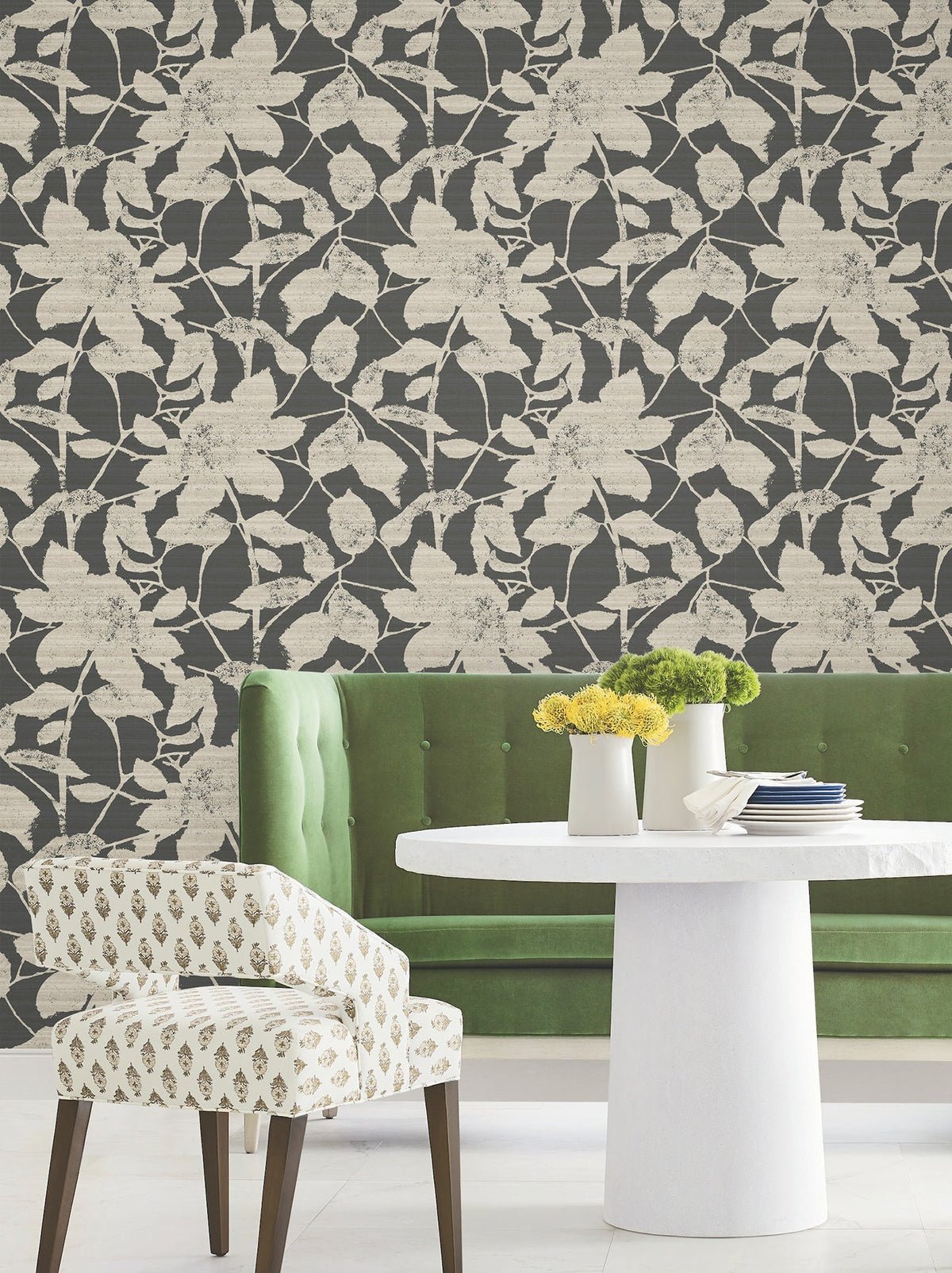 A modern dining area with a green tufted bench, white round table, patterned chair, and vases with yellow flowers. The background features York Wallcoverings’ Solange Botanical Wallpaper, showcasing large beige leaves and flowers on a dark backdrop.