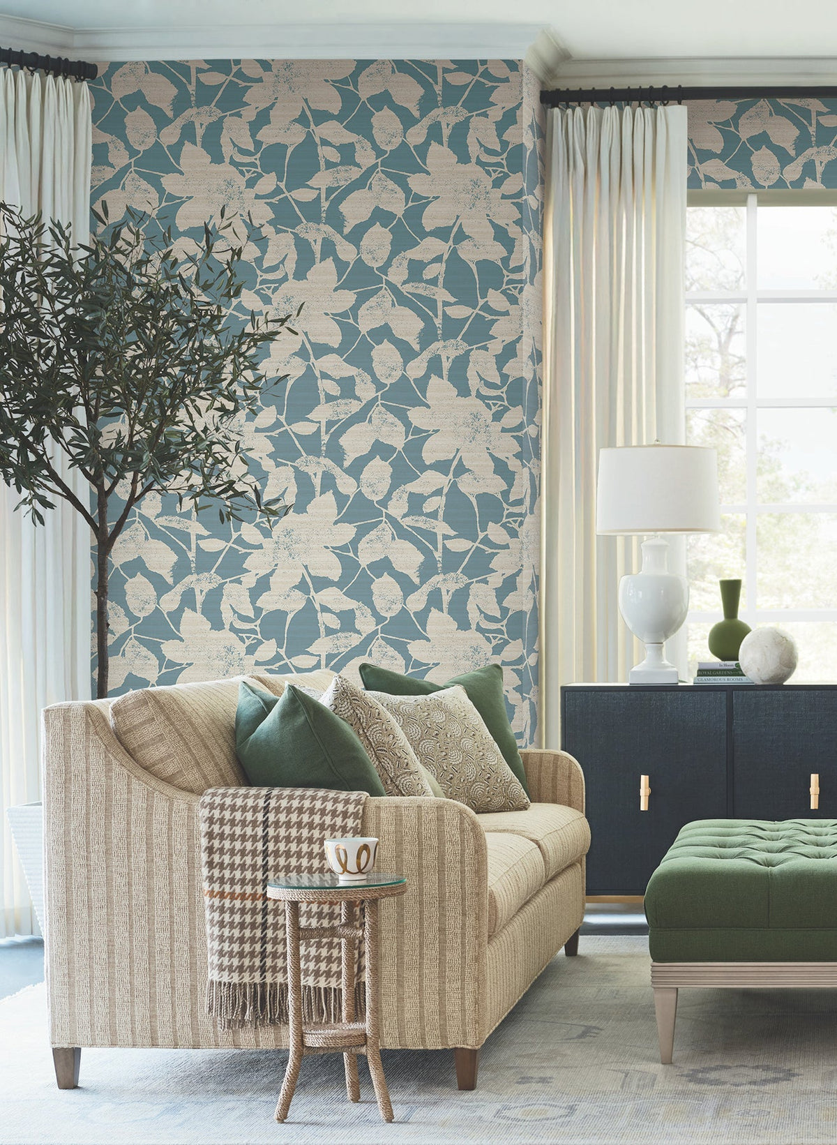 A cozy living room featuring a beige sofa with green pillows, a floral blue accent wall adorned with Solange Botanical Wallpaper by York Wallcoverings, a green ottoman, a dark cabinet, white lamp, large window with curtains, a small side table holding a mug, and a potted indoor tree.