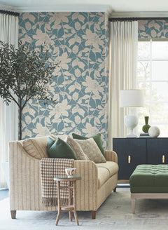 A cozy living room featuring a beige sofa with green pillows, a floral blue accent wall adorned with Solange Botanical Wallpaper by York Wallcoverings, a green ottoman, a dark cabinet, white lamp, large window with curtains, a small side table holding a mug, and a potted indoor tree.