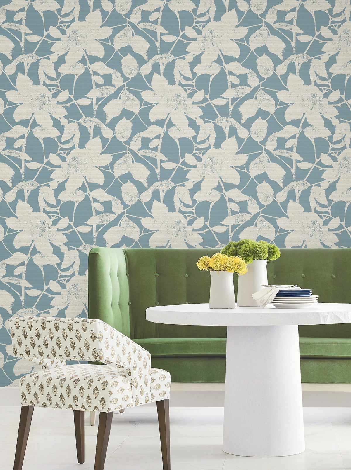 A green tufted bench, a white round table, and a patterned chair sit in front of a blue wall adorned with Solange Botanical Wallpaper by York Wallcoverings featuring a cream floral print. Yellow and green flowers in vases add a pop of color to the modern, airy room.
