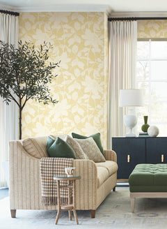 A cozy living room featuring a beige sofa with green and patterned pillows, a small side table with a mug, a tall potted tree, navy cabinet, white lamp, large window with white curtains, and York Wallcoverings’ Solange Botanical Wallpaper in yellow floral.