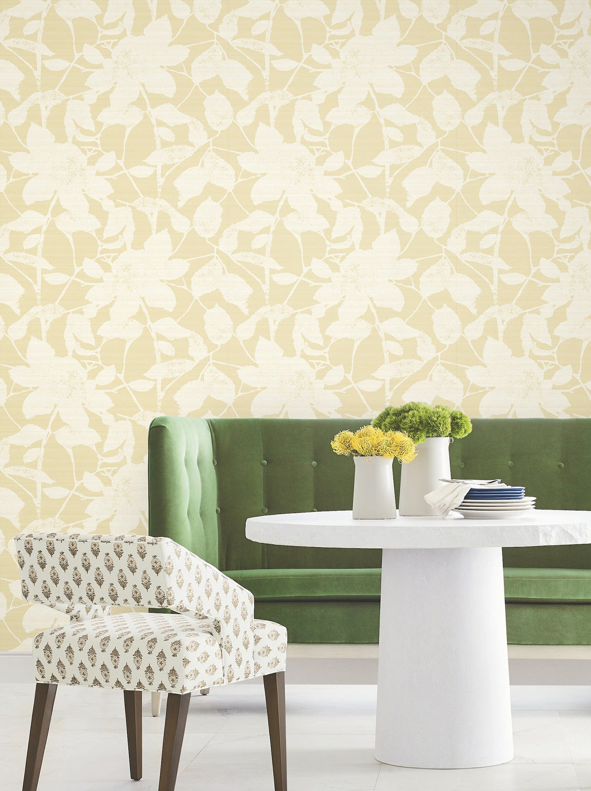 A green tufted sofa and a patterned chair surround a round white table with yellow and green flower arrangements. Solange Botanical Wallpaper by York Wallcoverings covers the wall behind them, adding a light yellow floral touch to the space.