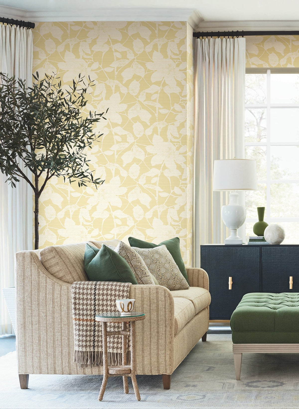 A cozy living room featuring a beige sofa with green and patterned pillows, a green ottoman, a side table topped with a mug, a potted tree, and natural light streaming through a window with cream curtains—all set against York Wallcoverings’ Solange Botanical Wallpaper.