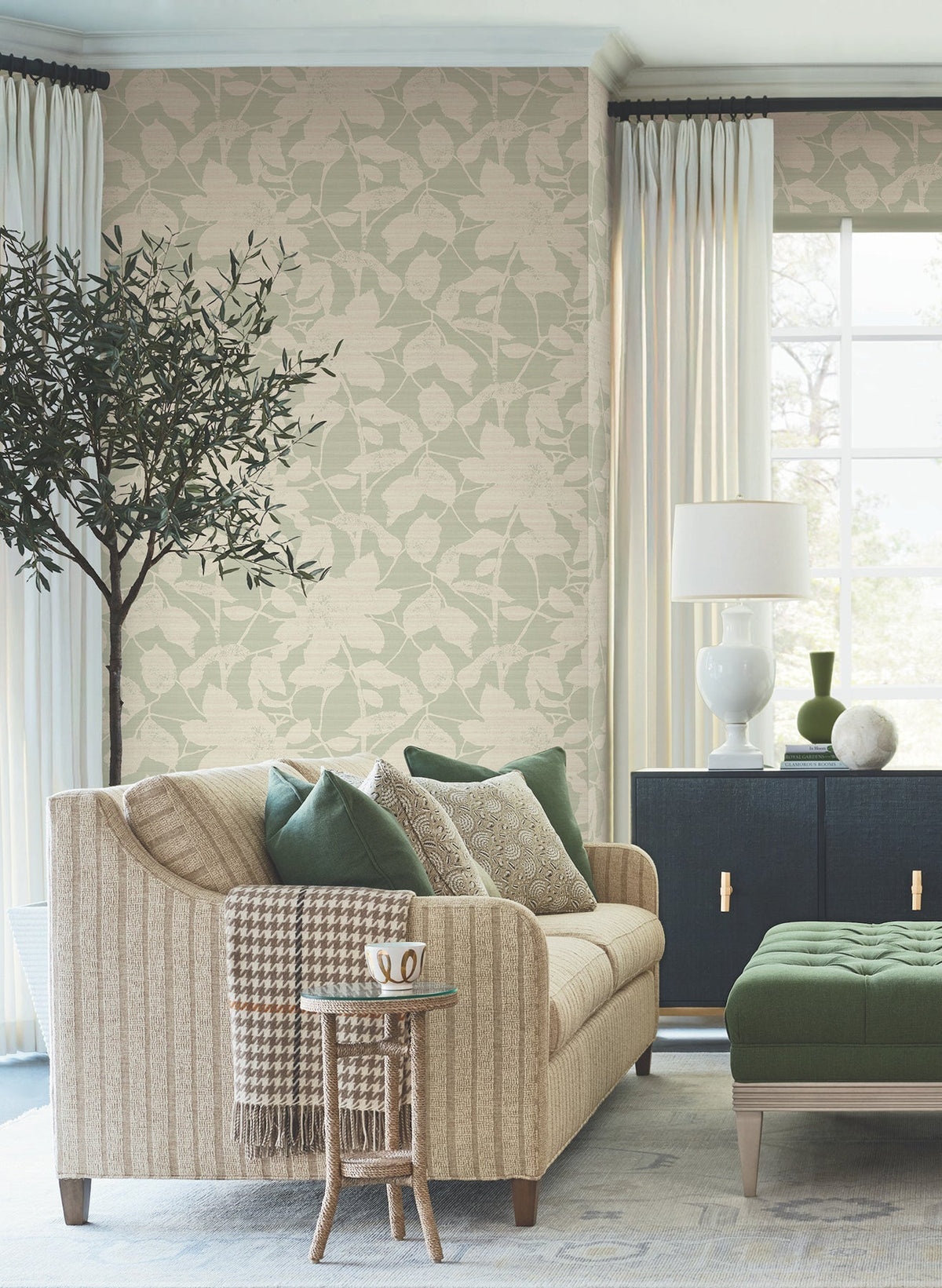 A cozy living room featuring Solange Botanical Wallpaper by York Wallcoverings, a beige sofa with green and patterned pillows, a small side table with a mug, a leafy tree, a black cabinet, a white lamp, and large windows adorned with cream curtains that let in natural light.