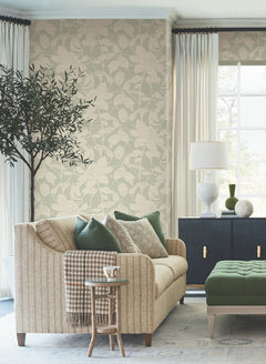 A cozy living room featuring Solange Botanical Wallpaper by York Wallcoverings, a beige sofa with green and patterned pillows, a small side table with a mug, a leafy tree, a black cabinet, a white lamp, and large windows adorned with cream curtains that let in natural light.