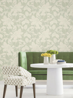 A green upholstered bench, a round white table with yellow flowers, and a patterned chair sit in front of York Wallcoverings’ Solange Botanical Wallpaper in a bright, modern room.
