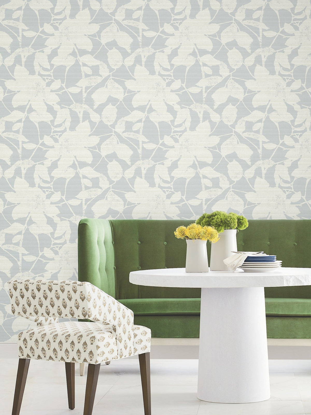 A green tufted sofa, round white table with vases of yellow flowers, and a patterned chair sit in front of light wallpaper featuring the Solange Botanical design by York Wallcoverings.
