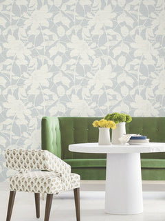 A green tufted sofa, round white table with vases of yellow flowers, and a patterned chair sit in front of light wallpaper featuring the Solange Botanical design by York Wallcoverings.