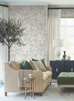 A stylish living room featuring a beige sofa with green cushions, a small side table holding a mug, a green ottoman, and a potted tree is complemented by cream curtains and natural light streaming through the window. The space is beautifully accented with Solange Botanical Wallpaper from York Wallcoverings.