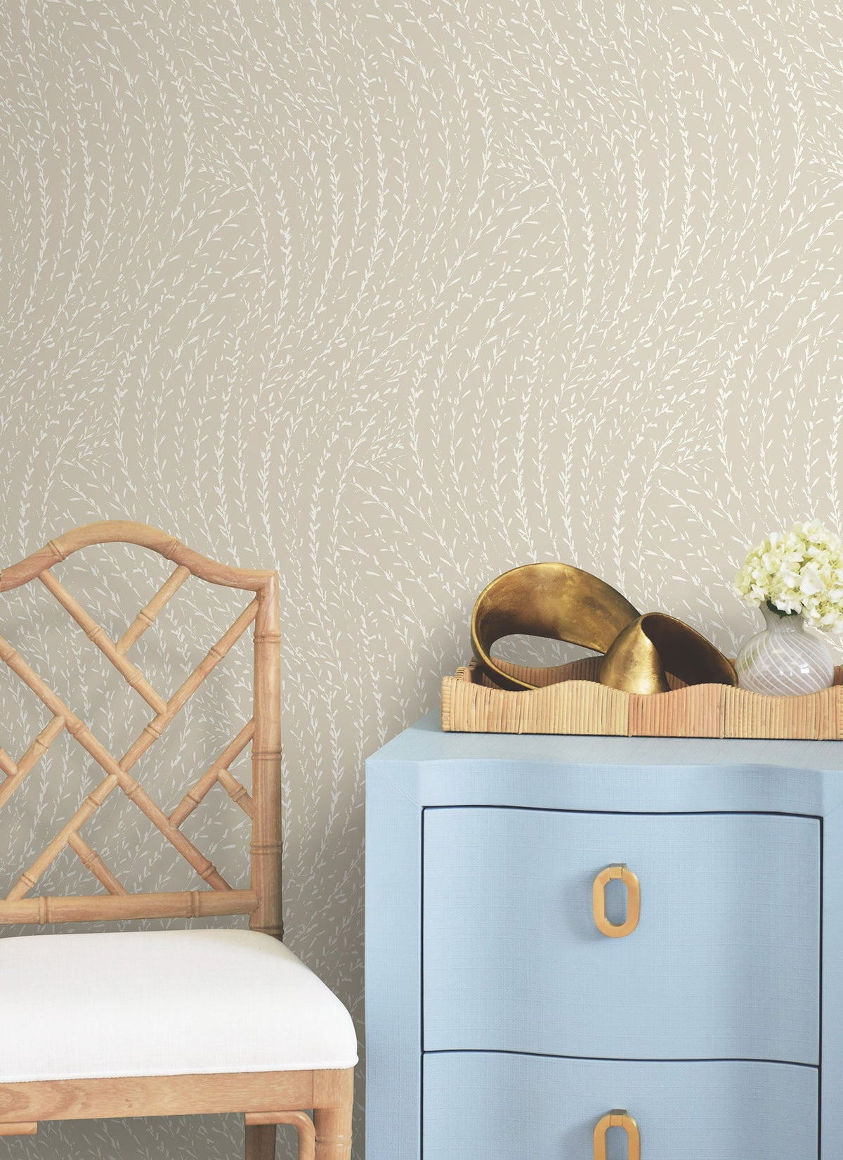 A light wooden chair sits next to a blue drawer with gold handles. On the drawer is a tray with gold decorative objects and a small bouquet of white flowers. The wall features York Wallcoverings’ Willow Song Botanical Wallpaper, adding a subtle beige leafy pattern to the space.