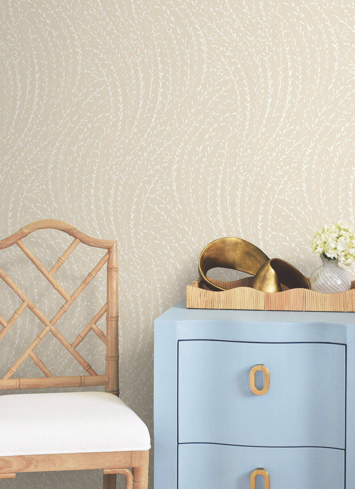 A light blue dresser with gold handles sits beside a bamboo chair. On the dresser is a tray with gold decor items and a small vase of white flowers. The wall features Willow Song Botanical Wallpaper by York Wallcoverings, showcasing a beige background with a subtle white leaf pattern.