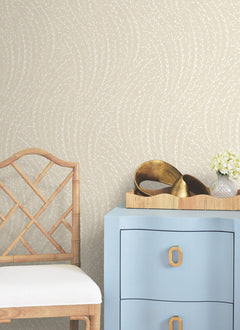 A light blue dresser with gold handles sits beside a bamboo chair. On the dresser is a tray with gold decor items and a small vase of white flowers. The wall features Willow Song Botanical Wallpaper by York Wallcoverings, showcasing a beige background with a subtle white leaf pattern.