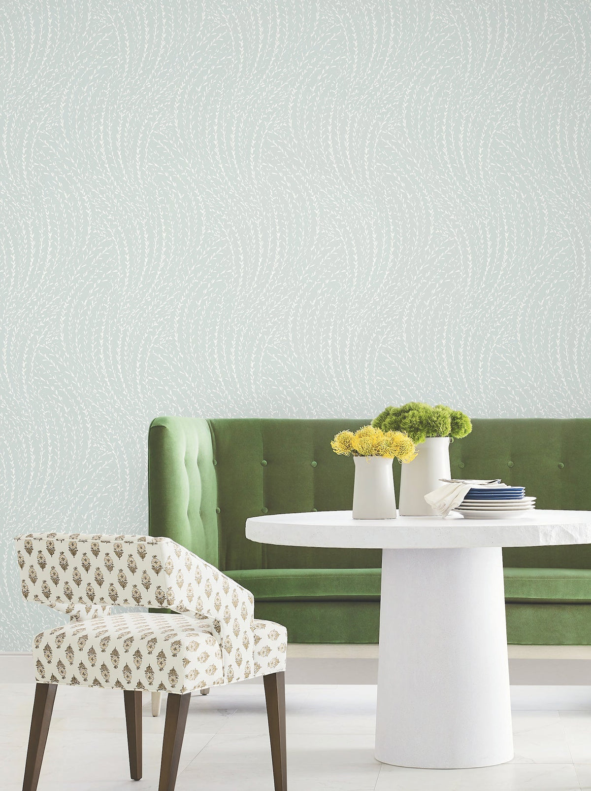 A green velvet bench, a white round table, and a patterned white chair sit in a room with pale Willow Song Botanical Wallpaper by York Wallcoverings. Three small vases with yellow and green flowers are on the table.
