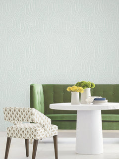 A green velvet bench, a white round table, and a patterned white chair sit in a room with pale Willow Song Botanical Wallpaper by York Wallcoverings. Three small vases with yellow and green flowers are on the table.