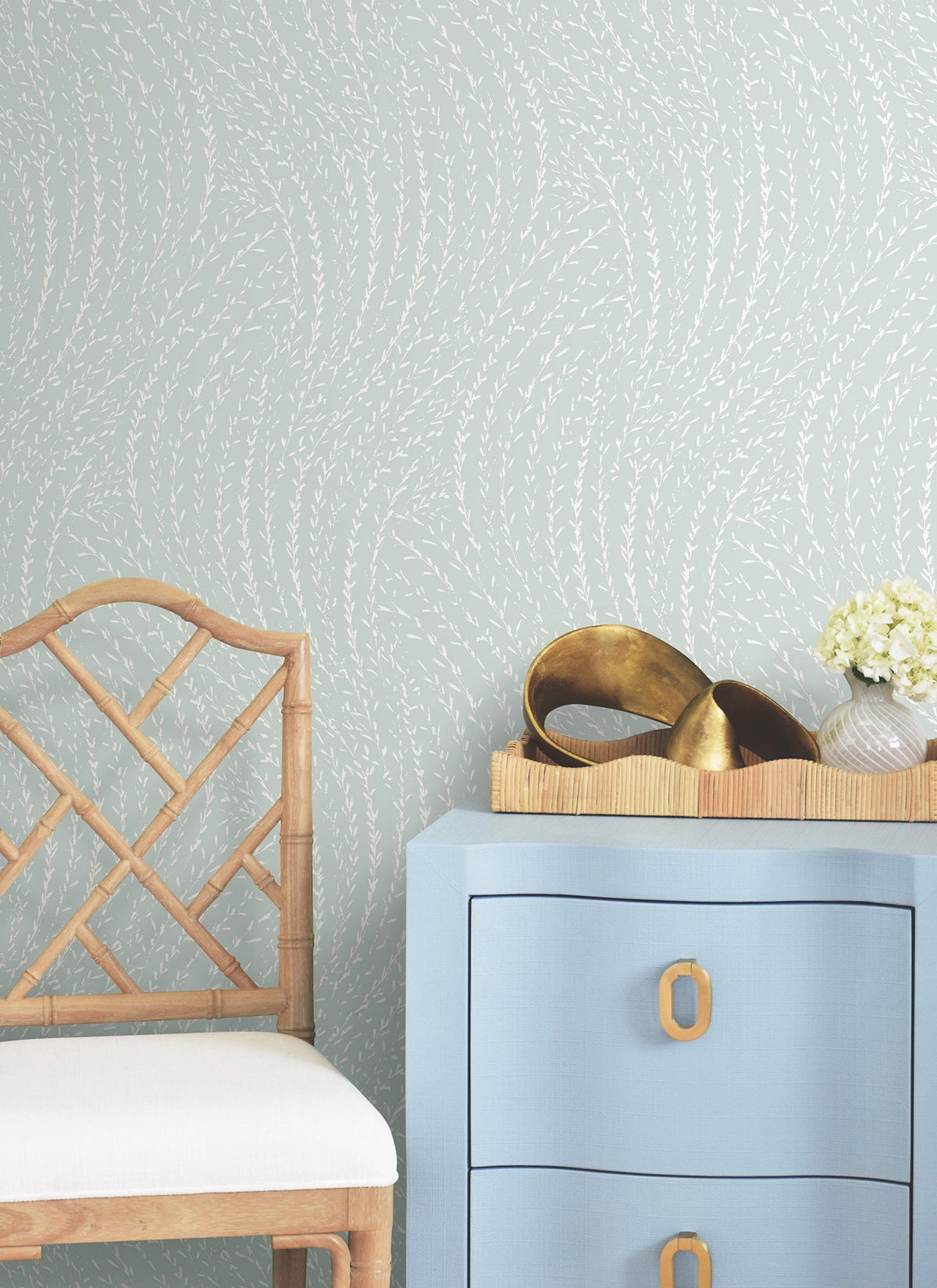 A wooden chair, a blue drawer with gold handles, a tray with brass décor, and a vase of white flowers are arranged against a wall adorned with York Wallcoverings’ Willow Song Botanical Wallpaper.