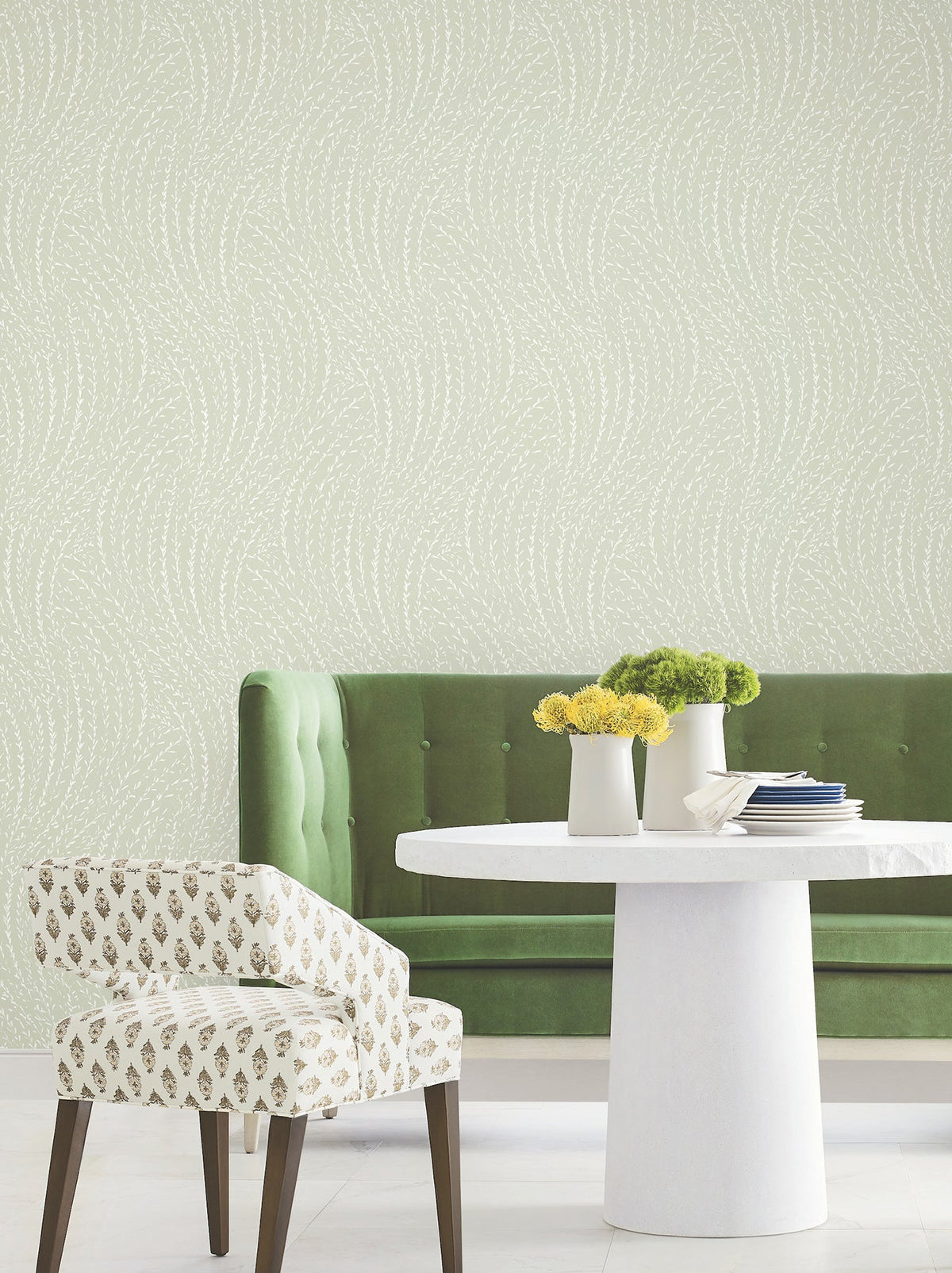 A green tufted bench and a patterned chair are positioned around a round white table adorned with vases of yellow and green flowers, set against Willow Song Botanical Wallpaper by York Wallcoverings, which features a subtle swirling dotted pattern.
