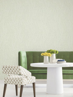 A modern dining area with a green tufted bench, white round table, patterned chair, and York Wallcoverings’ Willow Song Botanical Wallpaper, which features a subtle wavy dotted design. Vases with yellow and green flowers decorate the table.