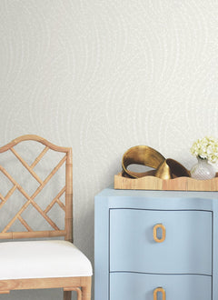 A bamboo chair with a white cushion sits next to a light blue dresser. On the dresser are a gold decorative object, a small white flower arrangement, and a beige tray. The background features Willow Song Botanical Wallpaper by York Wallcoverings.