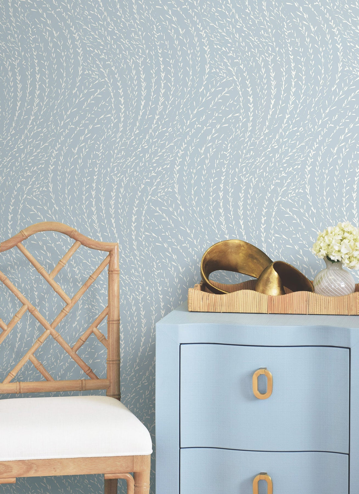 A light blue dresser with gold handles, a bamboo chair, a tray with gold decorative objects, and a small vase of white flowers are arranged against Willow Song Botanical Wallpaper by York Wallcoverings.