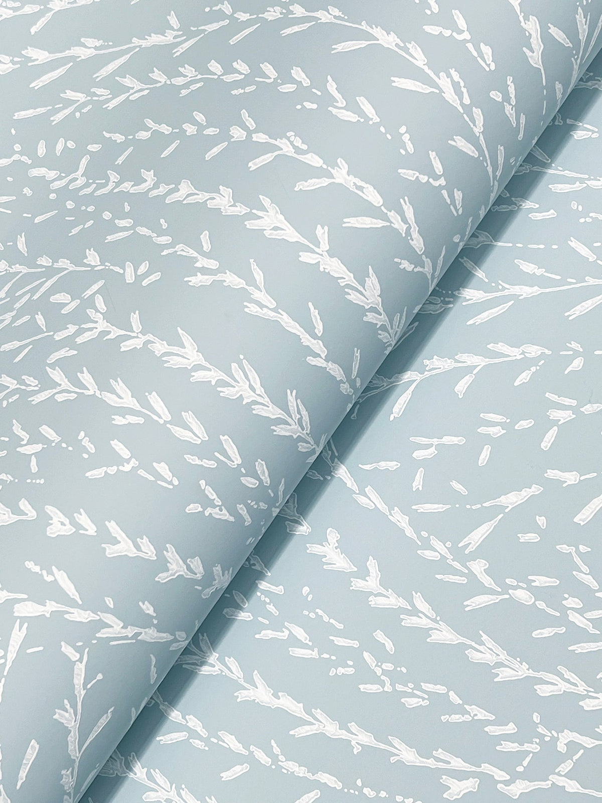 York Wallcoverings’ Willow Song Botanical Wallpaper features a light blue background adorned with a white, abstract leafy branch motif. The wallpaper displays a gently folded texture, highlighting its elegant repeating botanical design.