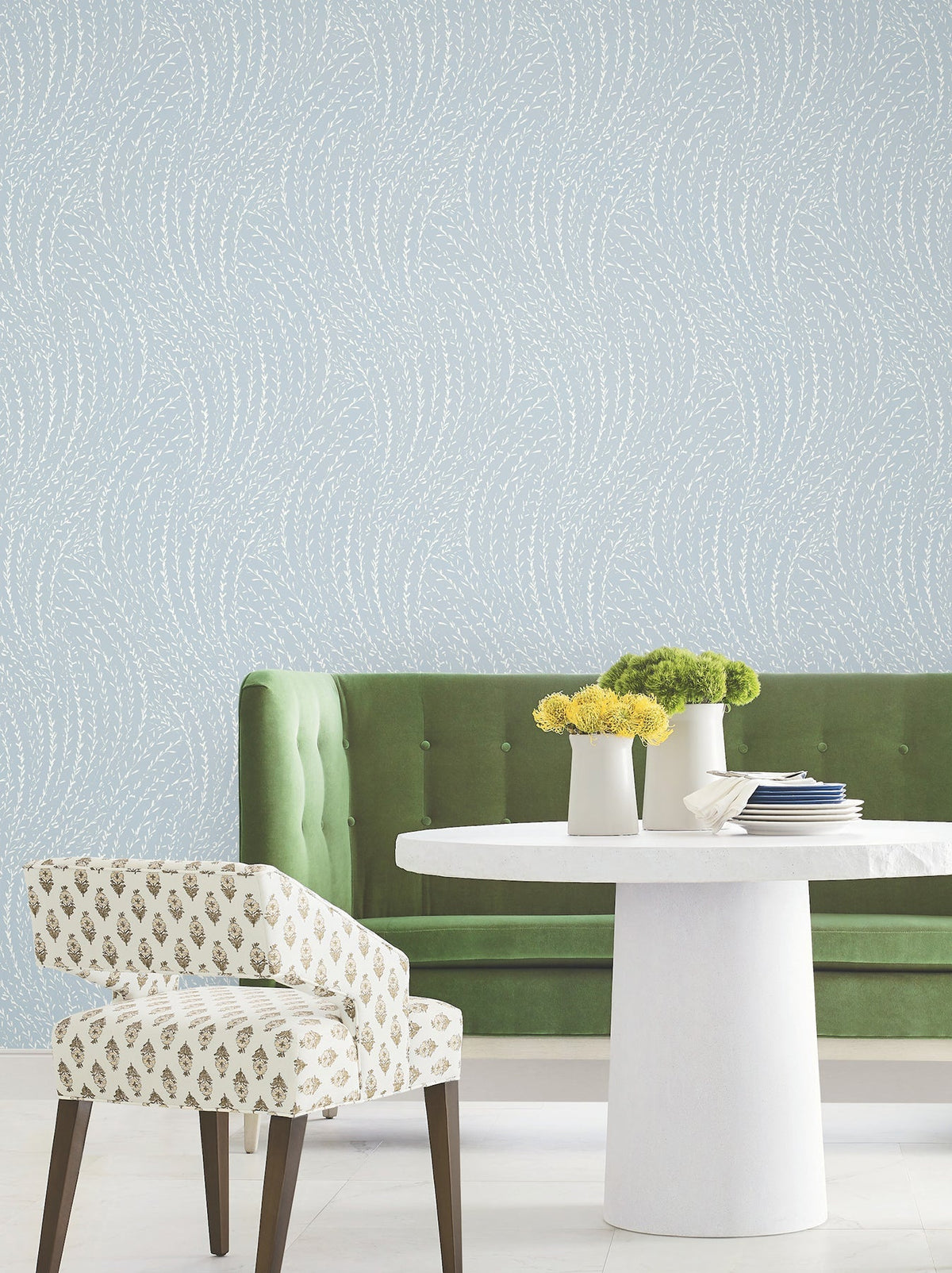 A green velvet sofa, a white round table with yellow flowers, and a patterned chair sit in front of Willow Song Botanical Wallpaper by York Wallcoverings, featuring a pale blue background with white wavy dotted lines.