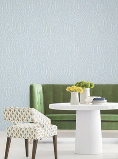 A green velvet sofa, a white round table with yellow flowers, and a patterned chair sit in front of Willow Song Botanical Wallpaper by York Wallcoverings, featuring a pale blue background with white wavy dotted lines.