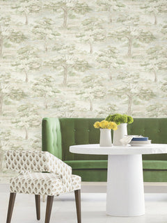 A round white table with yellow flowers, a patterned chair, and a green tufted bench sit in front of a wall adorned with the Moreno Woodland Wallpaper by York Wallcoverings, which features light green trees and a soft, nature-inspired pattern.