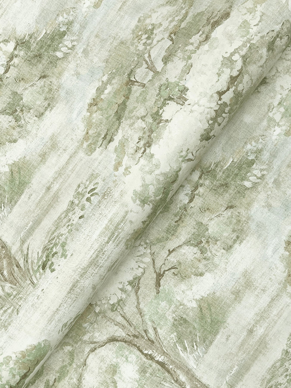 A close-up of York Wallcoverings’ Moreno Woodland Wallpaper reveals a nature-inspired design, featuring muted green trees and foliage printed on a light background. The wallpaper is slightly folded, highlighting its texture and the intricate pastoral scene.