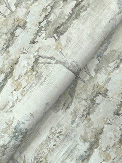 A close-up of Moreno Woodland Wallpaper by York Wallcoverings shows its delicate, muted pattern featuring trees with leafy branches in soft shades of gray, green, and cream. The wallpaper appears slightly folded, creating gentle shadows.