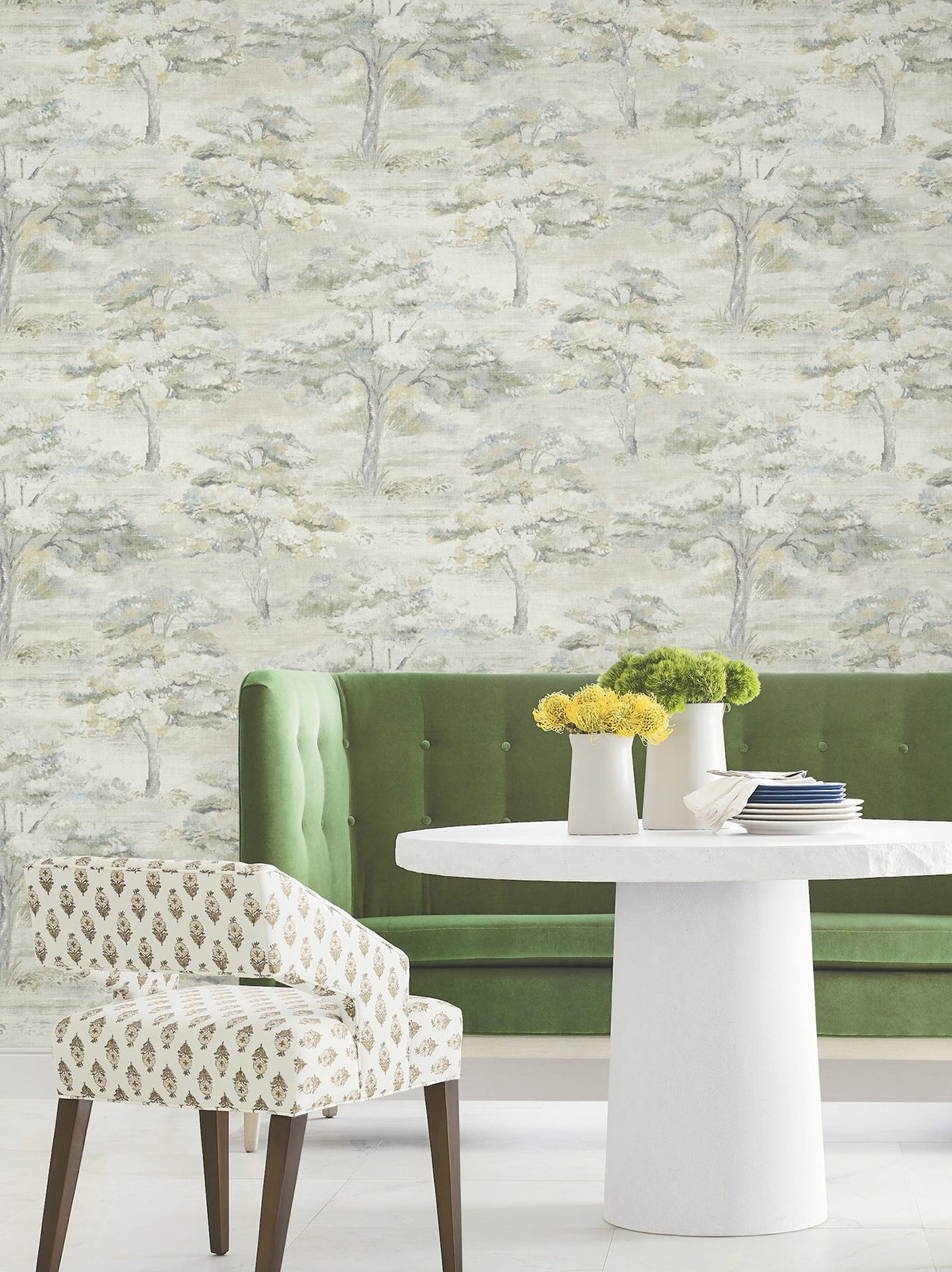 A green velvet sofa sits against a wall adorned with York Wallcoverings’ Moreno Woodland Wallpaper. In front, a round white table holds vases of yellow flowers. An upholstered chair with a small gold pattern stands nearby on a white floor.