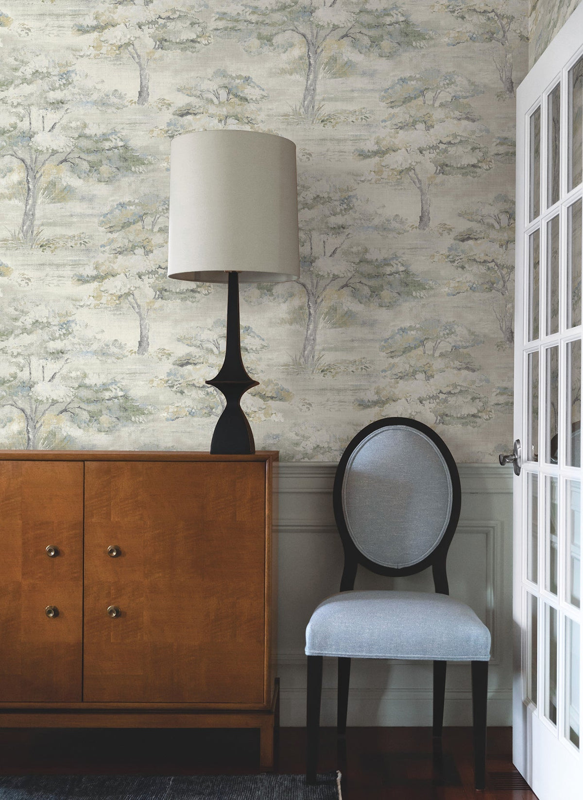 A wooden cabinet with a black lamp sits beside a light gray upholstered chair. The wall features Moreno Woodland Wallpaper by York Wallcoverings, and a glass-paned white door is partially visible on the right.