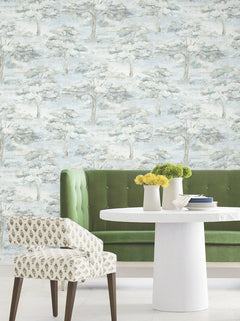 A green velvet sofa and a patterned chair are arranged by a round white table topped with yellow and green flowers, set against York Wallcoverings’ Moreno Woodland Wallpaper featuring a soft blue and gray tree landscape.