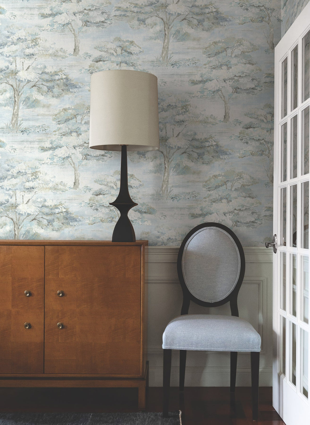 A wooden cabinet with a black lamp, a round-back gray chair, and French doors in a room adorned with Moreno Woodland Wallpaper by York Wallcoverings.