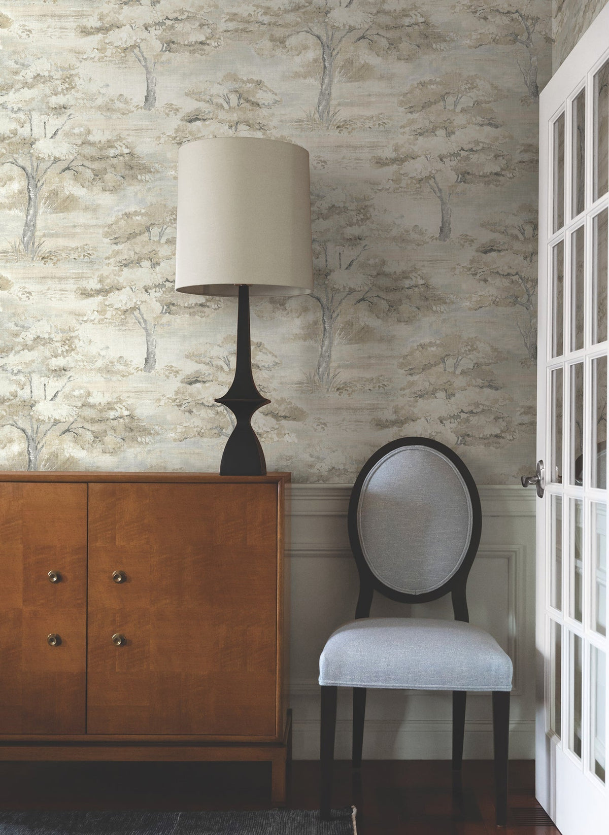 A wooden sideboard, a black lamp with a beige shade, and a gray cushioned chair are arranged against a wall adorned with Moreno Woodland Wallpaper by York Wallcoverings near a glass-paned door.