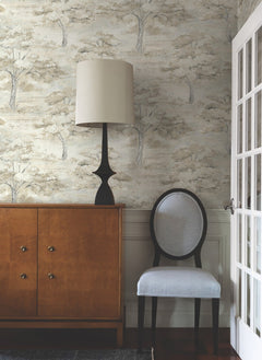A wooden sideboard, a black lamp with a beige shade, and a gray cushioned chair are arranged against a wall adorned with Moreno Woodland Wallpaper by York Wallcoverings near a glass-paned door.
