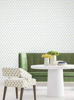 A green tufted bench, patterned chair, and round white table with vases of yellow flowers sit in front of a white wall adorned with Serene Villa Floral Stamp Wallpaper by York Wallcoverings.
